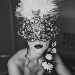 K&ouml;nnte beinhalten: Eine Frau tr&auml;gt eine schwarz-wei&szlig;e Maskenballmaske mit Federn und Strasssteinen. Sie tr&auml;gt ein schwarzes tr&auml;gerloses Kleid und hat einen Blumenschmuck im Haar.