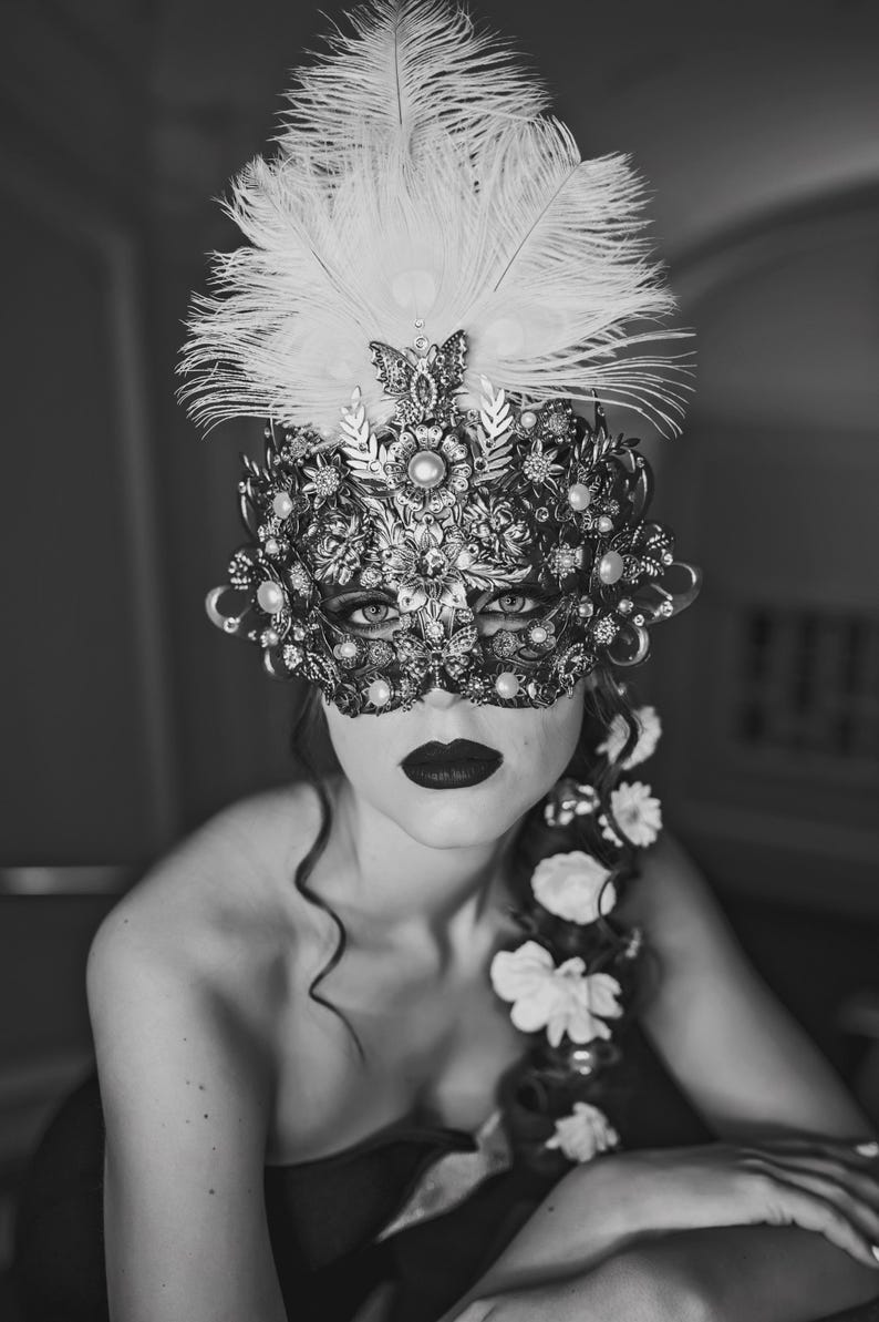 K&ouml;nnte beinhalten: Eine Frau tr&auml;gt eine schwarz-wei&szlig;e Maskenballmaske mit Federn und Strasssteinen. Sie tr&auml;gt ein schwarzes tr&auml;gerloses Kleid und hat einen Blumenschmuck im Haar.