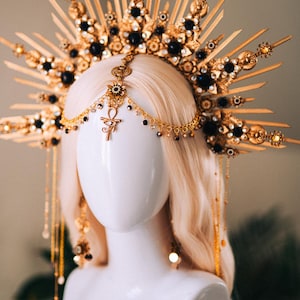 May include: A gold-toned headpiece with black accents and a sunburst design. The elaborate crown features intricate details, including black gemstones and dangling chains. The headpiece is displayed on a mannequin head with blonde hair, showcasing its ornate design.
