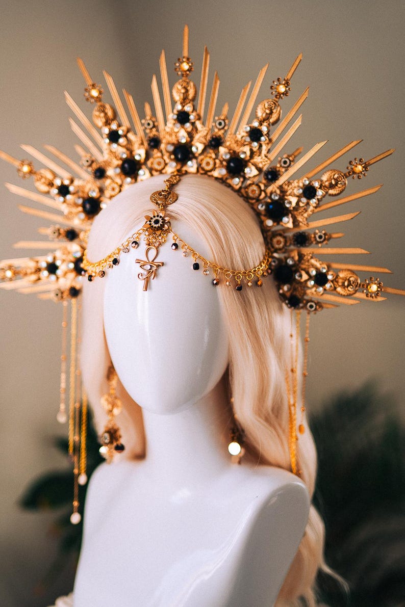 May include: A gold-toned headpiece with black accents and a sunburst design. The elaborate crown features intricate details, including black gemstones and dangling chains. The headpiece is displayed on a mannequin head with blonde hair, showcasing its ornate design.