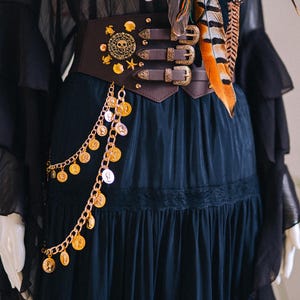 Puede incluir: Un cintur&oacute;n cors&eacute; marr&oacute;n oscuro con adornos dorados, que incluyen una calavera, estrellas y monedas, es el punto focal de esta imagen. El cintur&oacute;n tiene tres hebillas y se combina con un vestido negro y una bufanda con plumas naranjas y marrones.