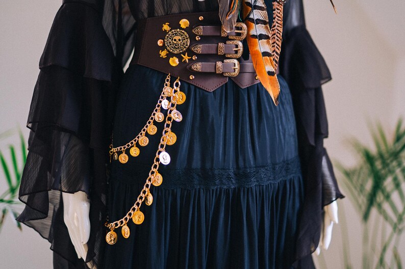 Puede incluir: Un cintur&oacute;n cors&eacute; marr&oacute;n oscuro con adornos dorados, que incluyen una calavera, estrellas y monedas, es el punto focal de esta imagen. El cintur&oacute;n tiene tres hebillas y se combina con un vestido negro y una bufanda con plumas naranjas y marrones.