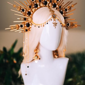 May include: A gold-toned headpiece with black accents and dangling chains, displayed on a white mannequin head. The elaborate design features ornate details and a central Ankh symbol, suggesting a celestial or regal theme. The headpiece is paired with matching earrings.