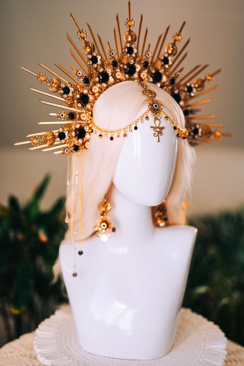 May include: A gold-toned headpiece with black accents and dangling chains, displayed on a white mannequin head. The elaborate design features ornate details and a central Ankh symbol, suggesting a celestial or regal theme. The headpiece is paired with matching earrings.