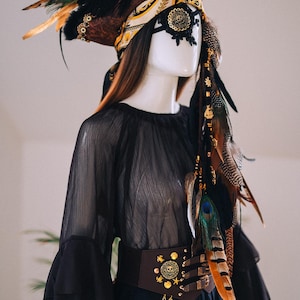 Puede incluir: Maniqu&iacute; con un conjunto negro de tem&aacute;tica pirata. El atuendo incluye un sombrero con plumas de pavo real, una m&aacute;scara de ojos negra, una blusa negra transparente y un cintur&oacute;n marr&oacute;n adornado con monedas de oro y detalles de calaveras. Una cadena decorativa cuelga del cintur&oacute;n.