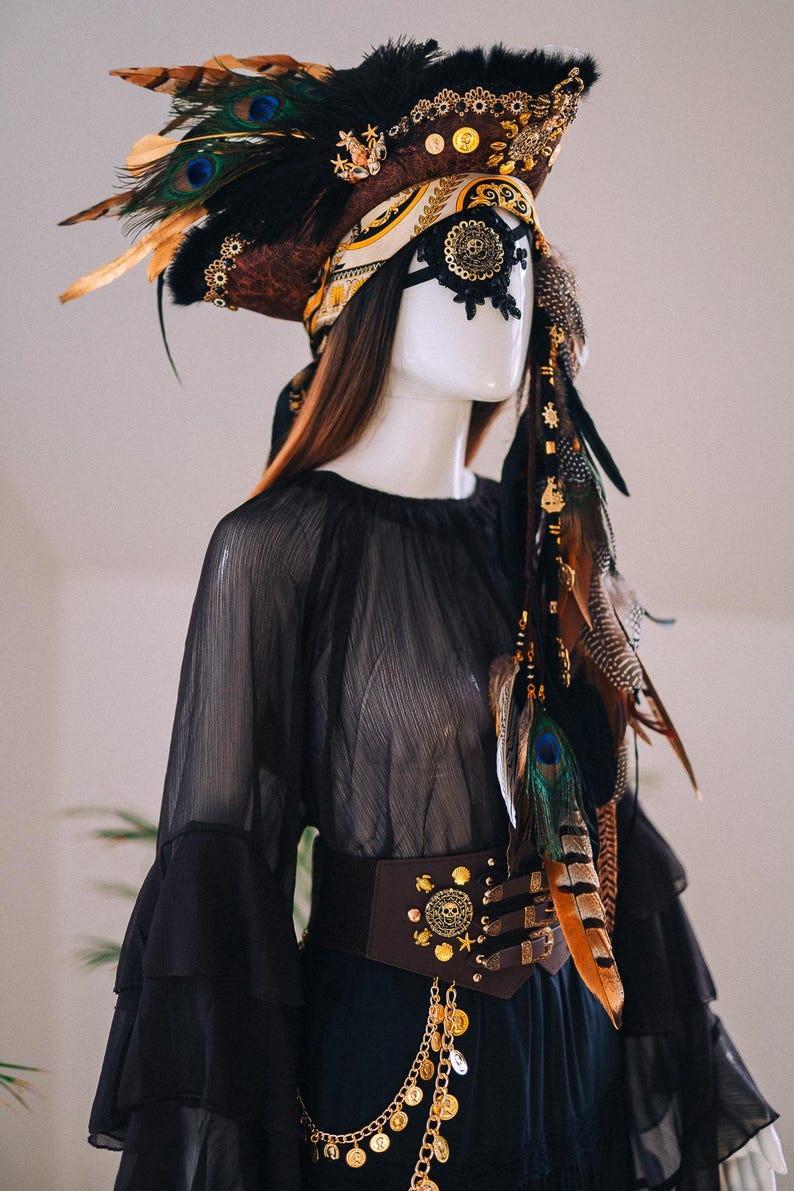 Puede incluir: Maniqu&iacute; con un conjunto negro de tem&aacute;tica pirata. El atuendo incluye un sombrero con plumas de pavo real, una m&aacute;scara de ojos negra, una blusa negra transparente y un cintur&oacute;n marr&oacute;n adornado con monedas de oro y detalles de calaveras. Una cadena decorativa cuelga del cintur&oacute;n.