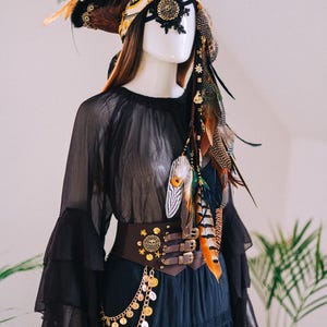 Puede incluir: Maniqu&iacute; vestido con un conjunto negro de tem&aacute;tica pirata. El atuendo incluye un tocado de plumas con detalles dorados, un parche decorativo en el ojo y un cintur&oacute;n marr&oacute;n con monedas de oro. El conjunto se completa con una blusa negra transparente.