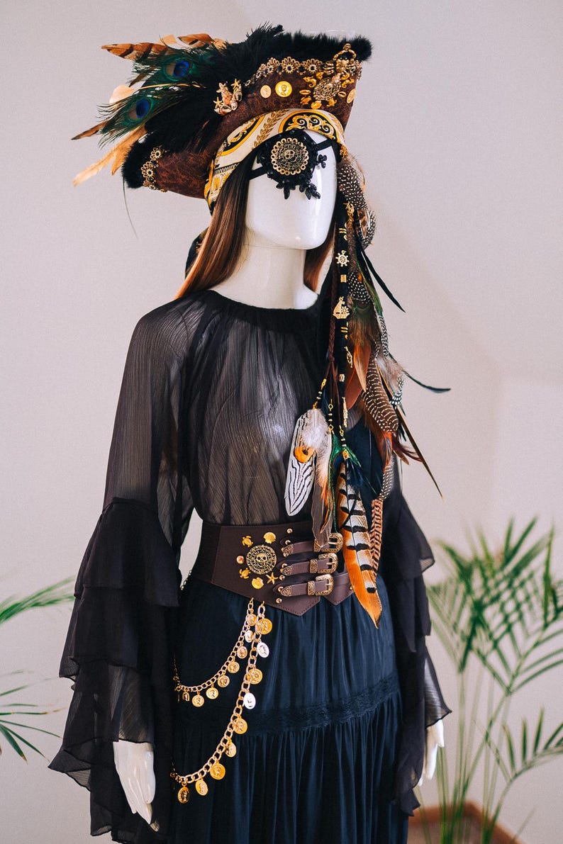 Puede incluir: Maniqu&iacute; vestido con un conjunto negro de tem&aacute;tica pirata. El atuendo incluye un tocado de plumas con detalles dorados, un parche decorativo en el ojo y un cintur&oacute;n marr&oacute;n con monedas de oro. El conjunto se completa con una blusa negra transparente.
