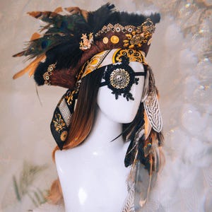 Puede incluir: Tocado decorativo en tonos marr&oacute;n, negro y dorado. Presenta plumas, un parche en el ojo con forma de calavera y detalles ornamentados. El dise&ntilde;o incluye una mezcla de texturas y patrones, creando un aspecto &uacute;nico y art&iacute;stico.