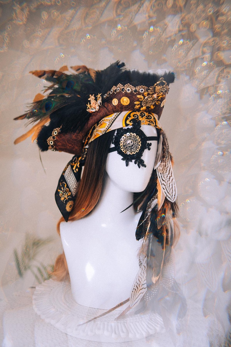 Puede incluir: Tocado decorativo en tonos marr&oacute;n, negro y dorado. Presenta plumas, un parche en el ojo con forma de calavera y detalles ornamentados. El dise&ntilde;o incluye una mezcla de texturas y patrones, creando un aspecto &uacute;nico y art&iacute;stico.