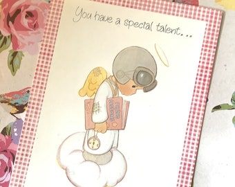 May include: A greeting card with a pink and white gingham border. The card features a cartoon angel wearing a pilot's helmet and holding a book that says "Flying Lessons". The text on the card reads "You have a special talent..."