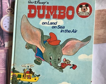 May include: A vintage children's book cover featuring Dumbo the elephant flying in the air with Timothy Mouse. The book title is "Walt Disney's Dumbo on Land on Sea in the Air" and the cover is illustrated with a blue sky, a yellow helicopter, a red and white ship, and a circus tent.