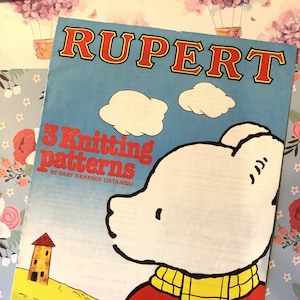 May include: A vintage Rupert Bear knitting pattern booklet featuring three knitting patterns. The cover features a cartoon illustration of Rupert Bear wearing a red jumper and yellow scarf. The title of the booklet is "3 Knitting patterns by Gary Kennedy (Intarsia)".