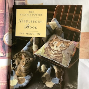 May include: A book titled "The Beatrix Potter Needlepoint Book" by Pat Menchini. The cover features a brown and white cat sitting on a brown chair with a needlepoint pillow featuring a cat. The chair is covered in a blue, brown, and white knitted blanket.
