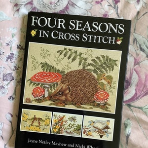 May include: A book titled "Four Seasons in Cross Stitch" with a black cover and colourful cross stitch designs of a hedgehog, a rabbit, flowers, and birds. The book is by Jayne Netley Mayhew and Nicki Wheeler.
