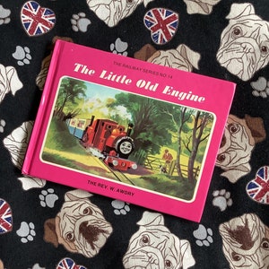 Könnte beinhalten: Ein rosa Buch mit dem Titel "The Little Old Engine" aus "The Railway Series No. 14" von The Rev. W. Awdry. Der Umschlag zeigt eine rote Lokomotive mit einem lächelnden Gesicht.