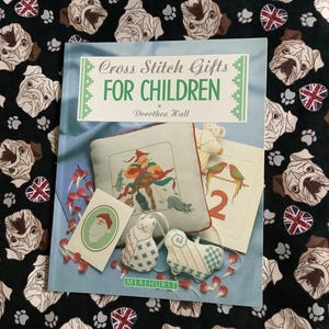 May include: A craft book titled "Cross Stitch Gifts For Children" by Dorothea Hall, featuring cross-stitch projects. The book rests on black fabric patterned with pug faces, paw prints, and Union Jack hearts.