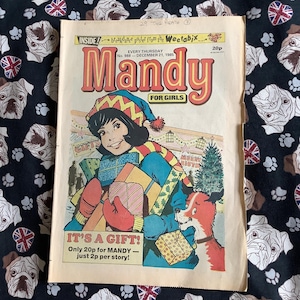 May include: A vintage Mandy comic book from 21 December 1985, featuring a colourful cover illustration of a person holding wrapped Christmas gifts alongside a dog. The text includes Mandy For Girls, It's A Gift, and Weetabix breakdance poster.