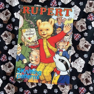 May include: A vintage "Rupert" comic book featuring a bear character in a red shirt and yellow pants, holding a smaller book. The cover text reads "5 Knitting Patterns". The background is a black fabric with pug dog and Union Jack patterns.