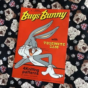 Może przedstawiać: Czerwono-biała książeczka z wzorami na robótki na drutach z postaciami Bugs Bunny i Yosemite Sam. Na okładce znajduje się kreskówkowa ilustracja Bugs Bunny'a z tekstem "Bugs Bunny with Yosemite Sam" i "4 Knitting Patterns for children and adults".