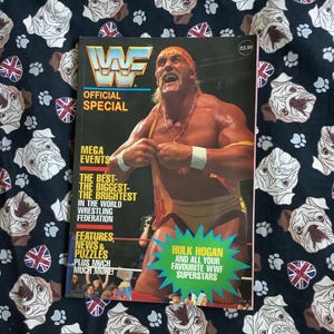 Könnte beinhalten: Ein Vintage WWF Official Special Magazin mit Hulk Hogan auf dem Cover. Das Magazin hat ein blaues, rotes und gelbes Farbschema mit Text, der "Mega Events" und "Features, News & Puzzles" lautet. Das Magazin liegt auf einem schwarzen Stoff mit einem Muster aus weißen Bulldoggen-Gesichtern und Union Jack-Flaggen.