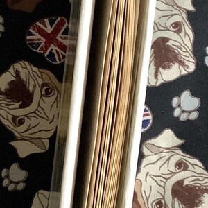 Puede incluir: Un &aacute;lbum de fotos abierto, con lomo blanco, mostrando las p&aacute;ginas. El fondo es una tela negra estampada con caras de bulldog, huellas de patas y emblemas de la Union Jack. El &aacute;lbum est&aacute; abierto.