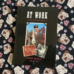 Pode incluir: Um livro preto intitulado "AT WORK" com uma foto de um homem de uniforme. O livro inclui o texto "BYGONE BRITAIN 1900-1970". O livro está colocado sobre um tecido preto decorado com rostos de buldogues, pegadas e bandeiras Union Jack.