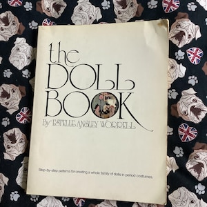 May include: A vintage book titled "the DOLL BOOK" by Estelle Ansley Worrell. The cover is cream-colored with black text and a small illustration. The background is a dark fabric patterned with pug faces, paw prints, and Union Jack hearts.
