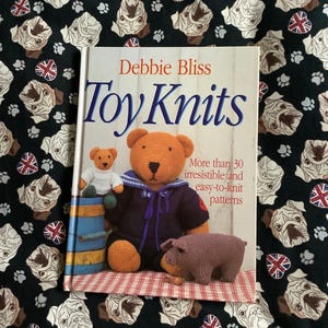 May include: A book titled "Toy Knits" by Debbie Bliss, featuring a knitted teddy bear, pig, and other toys. The cover art includes the text "More than 30 irresistible and easy-to-knit patterns." The background is a fabric with pug faces and Union Jack flags.