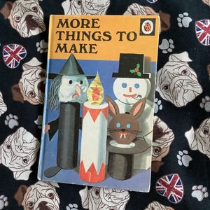 以下が含まれることがあります： 「MORE THINGS TO MAKE」というタイトルのヴィンテージ子供向けの本。魔女、ろうそく、雪だるま、ウサギのイラストが描かれています。本は黄色い表紙に青と白の文字が書かれています。背景は、ブルドッグの顔、足跡、ユニオンジャックのハートが描かれた濃い色の生地です。