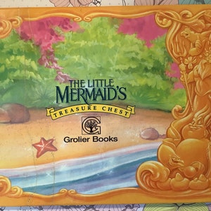 Pu&ograve; includere: Una copertina di libro gialla, arancione e verde con il titolo "Il forziere del tesoro della Sirenetta" e il logo Grolier Books. La copertina presenta un'illustrazione di cartone animato di una sirena e un cavalluccio marino.