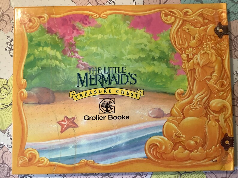 Pu&ograve; includere: Una copertina di libro gialla, arancione e verde con il titolo "Il forziere del tesoro della Sirenetta" e il logo Grolier Books. La copertina presenta un'illustrazione di cartone animato di una sirena e un cavalluccio marino.