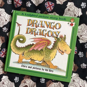 May include: A children's book titled "Drago Dragon" with a green cover and a cartoon dragon illustration. The book is a Ladybird picture story book, with text by Val Biro. The background is a black fabric with a pattern of bulldog faces and Union Jack flags.
