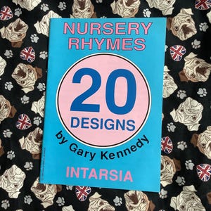 Könnte beinhalten: Ein blaues und rosa Buch mit dem Titel "Nursery Rhymes" mit einem rosa Kreis, der die Zahl "20" und den Text "Designs by Gary Kennedy Intarsia" enthält.