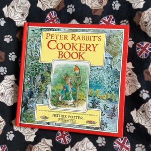 Peut inclure: Un livre de cuisine "Peter Rabbit" à couverture rigide avec une bordure rouge. La couverture présente une illustration de Pierre Lapin et d'un oiseau. Le livre est posé sur un fond noir avec un motif de bouledogues, d'empreintes de pattes et de drapeaux britanniques.