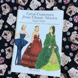 May include: A book titled "Great Costumes from Classic Movies: Paper Dolls" with a white background and three paper dolls in colorful dresses. The book cover features the text "30 Fashions by Adrian, Edith Head, Walter Plunkett and Others" and "Tom Tierney".