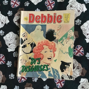 以下が含まれることがあります： 「Debbie」というタイトルのヴィンテージコミック。表紙には「Di's Disguises」の文字と、様々な衣装を着た女性たちのイラストが描かれています。背景は、ブルドッグの顔とユニオンジャックのエンブレムが描かれた黒い生地です。