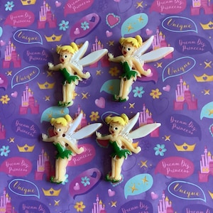 May include: Four fairy figurines with blonde hair, green dresses, and white wings are displayed on a purple background. The background has castles, crowns, hearts, and speech bubbles with the text "Dream Big Princess" and "Unique".