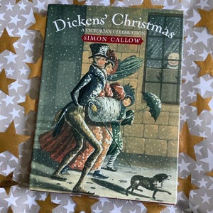以下が含まれることがあります： サイモン・カロウ著「ディケンズのクリスマス：ビクトリア朝の祝典」というタイトルの本。表紙には、当時の衣装を着た人物、犬、雪が降るビクトリア朝のシーンが描かれています。本は、金の星が散りばめられた白い布の上にあります。