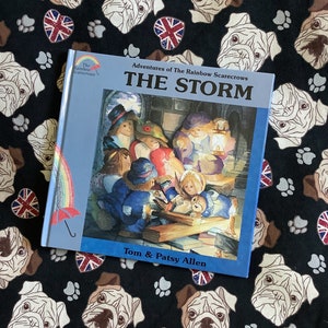May include: A blue book with the title "The Storm" and the subtitle "Adventures of The Rainbow Scarecrows" by Tom & Patsy Allen. The cover features an illustration of a group of scarecrows gathered around a table in a dimly lit room.