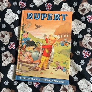 May include: A vintage Rupert Bear book with an orange cover and the title "Rupert" in large, bold, white letters. The cover features an illustration of Rupert Bear in a red and white checked shirt, standing on a wooden cart with a squirrel and a mouse. The book's title is "The Daily Express Annual".