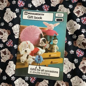 May include: A Woolworth gift book with the number 503 on the cover, featuring knitted toy patterns. The cover shows a pink knitted cushion, a pig, a dog, a snowman, and other knitted toys. The background is a black fabric with a pattern of bulldog faces and Union Jack flags.