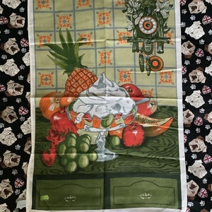 May include: A green and white tea towel with a fruit salad design. The design features a pineapple, grapes, apples, oranges, and a bowl of whipped cream. A cuckoo clock is in the top right corner of the towel.