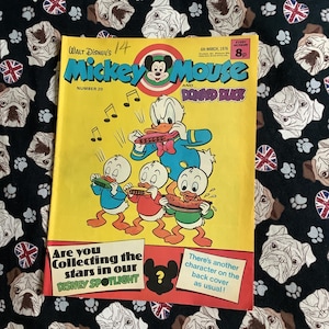 Könnte beinhalten: Ein Vintage-Comic von Mickey Mouse und Donald Duck, Nummer 20, mit einem gelben Cover mit Zeichentrickfiguren. Das Cover enthält Text und Illustrationen der Charaktere, die Instrumente spielen. Der Hintergrund ist ein schwarzer Stoff mit einem Muster aus Bulldoggen-Gesichtern und Pfotenabdrücken.