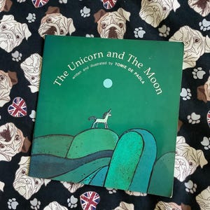 以下が含まれることがあります： トミー・デ・ポーラ著の絵本「The Unicorn and The Moon」。表紙には、ユニコーンと月が描かれた緑色の風景が描かれています。ブルドッグの顔、足跡、ユニオンジャックのエンブレムがパターン化された黒い生地の上に本が置かれています。
