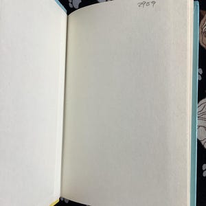 May include: Open book with blank pages. The book's spine is light blue, and the pages are off-white. The background is a black fabric with a pattern of dog faces and paw prints. The number "2909" is written in pencil on the top right page.