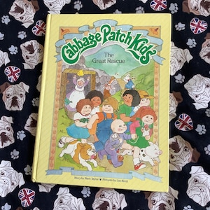 Könnte beinhalten: Ein gelbes Buch mit einem grünen Titel, der "Cabbage Patch Kids The Great Rescue" lautet. Der Umschlag zeigt eine Illustration der Cabbage Patch Kids-Charaktere, darunter eine Babypuppe, ein Hund und ein Schwein.