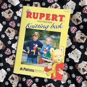 Può includere: Un libro di maglia giallo con il titolo "The Rupert and Friends Knitting Book" che presenta un'immagine di Rupert Bear e due bambini. Il libro è su un tessuto nero con un motivo di carlini e impronte di zampe.