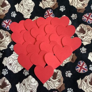 May include: A collection of red heart-shaped cutouts layered over a black fabric background featuring a repeating pattern of pug faces, grey paw prints, and small Union Jack heart icons. The red hearts are solid, flat, and have stitched edges.