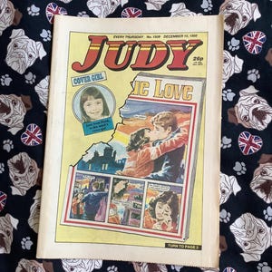 以下が含まれることがあります： 1988年12月10日発行の雑誌「Judy」のヴィンテージ版。表紙にはカバーガールと「IC Love」というタイトルの漫画が掲載されています。雑誌は黄色と赤の見出しで、白いブルドッグとユニオンジャックのパターンが施された黒い生地の上に置かれています。